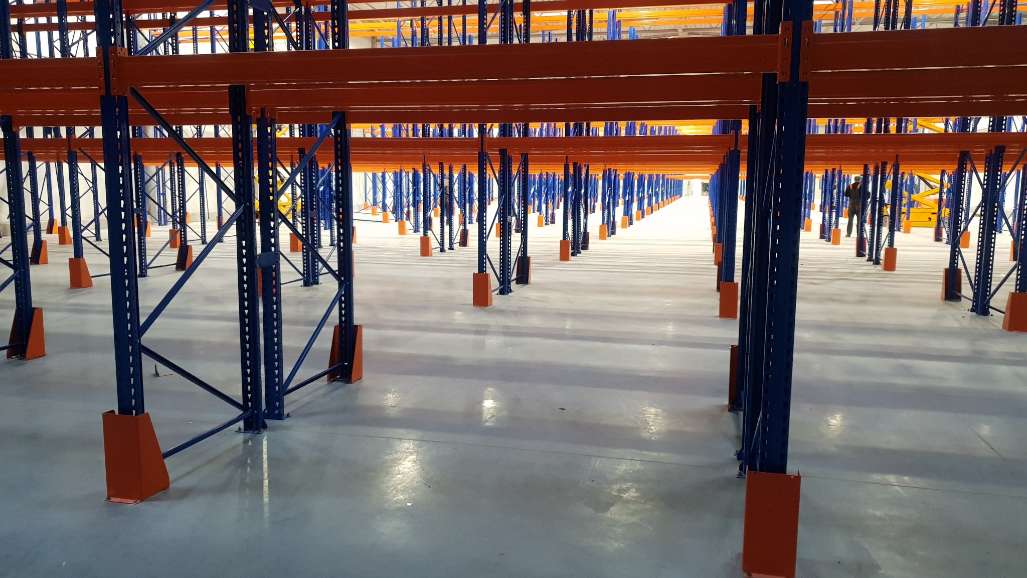 Długi rząd niebieskich metalowych słupków z pomarańczowymi podstawami i poprzeczkami, tworzących system regałów w pustym magazynie z błyszczącą podłogą.