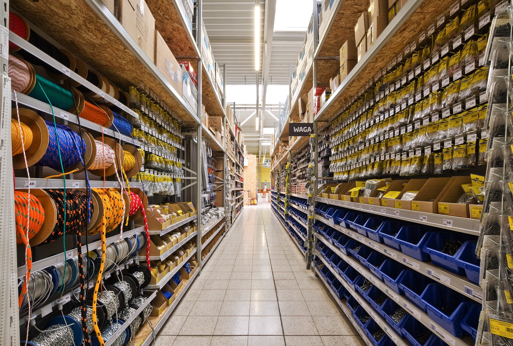 Długi kadr z perspektywy podłogi na alejkę w sklepie z artykułami budowlanymi, gdzie na metalowych regałach z drewnianymi półkami ułożone są szpule kolorowych lin, opakowania z elementami złącznymi...