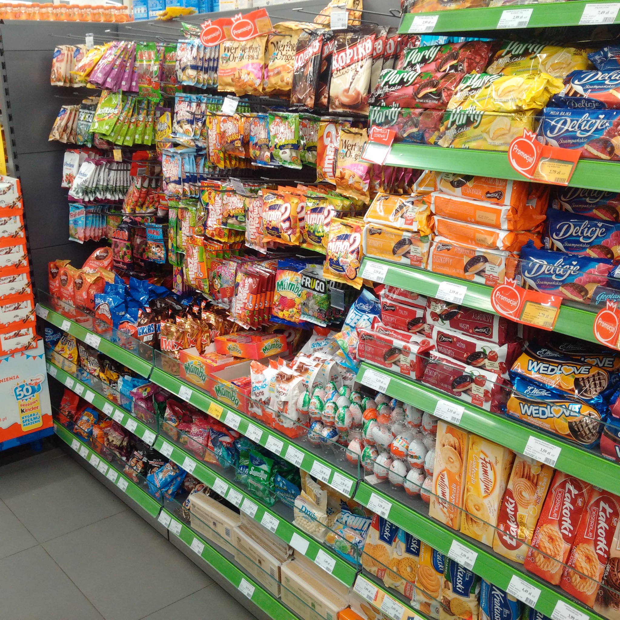 Półki sklepowe wypełnione różnorodnymi słodyczami: batoniki, ciastka, cukierki w opakowaniach, z widocznymi cenami i etykietami promocyjnymi.
