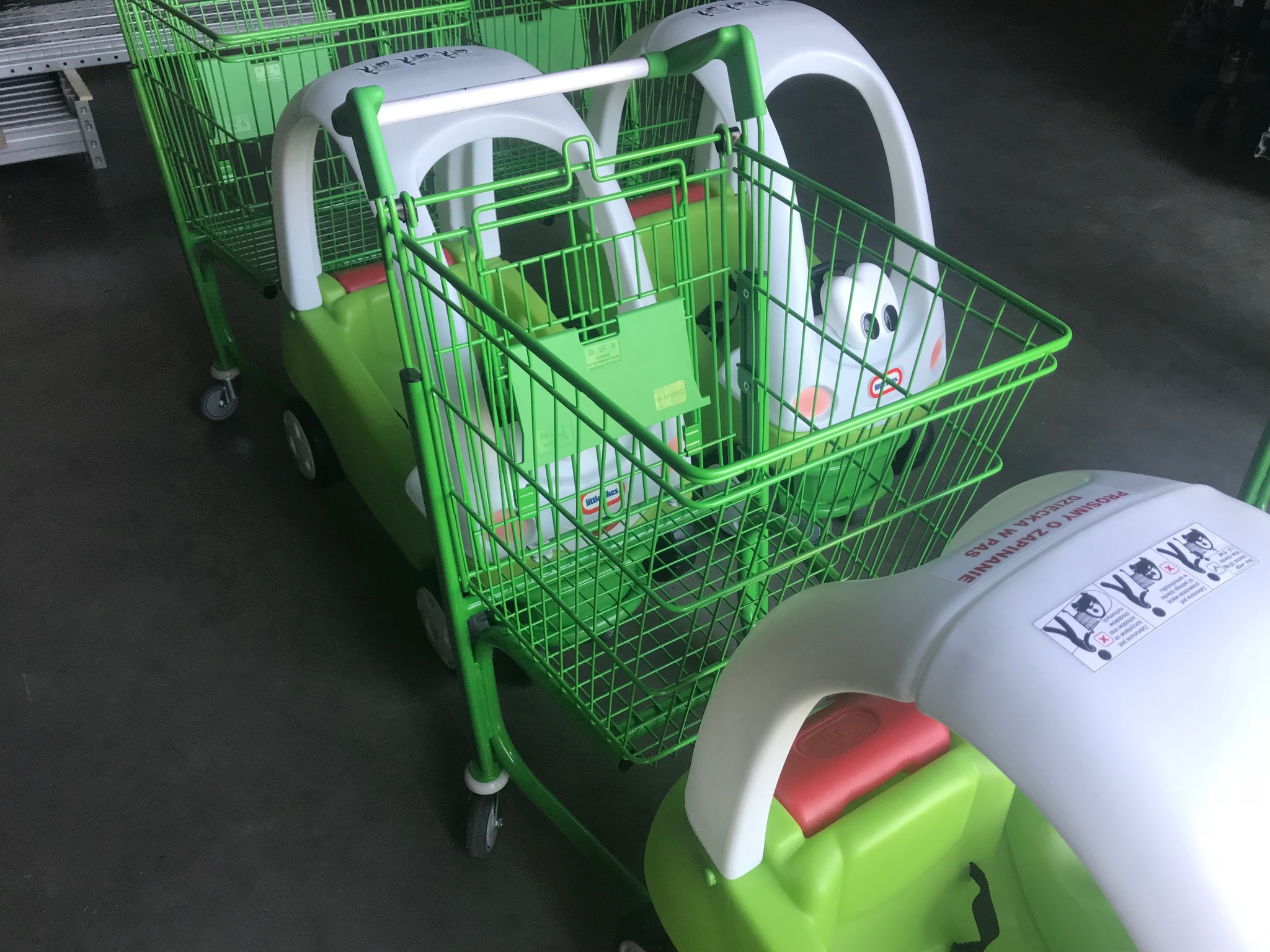 Szereg zielonych wózków sklepowych dla dzieci z plastikową obudową w kształcie samochodu i naklejką z instrukcją bezpieczeństwa.