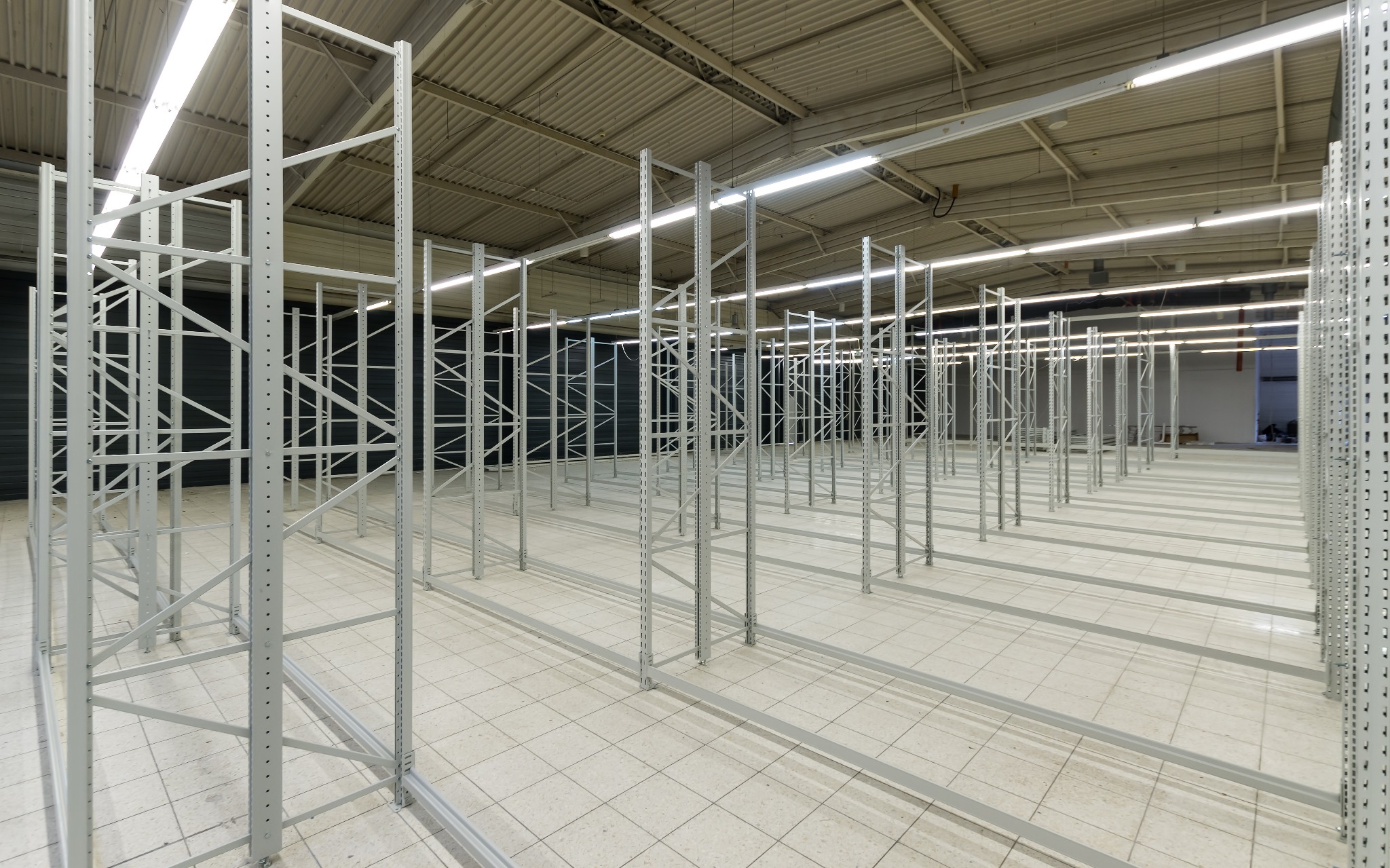Metalowe ramy regałów magazynowych ustawione w rzędach na jasnej podłodze w magazynie, widoczne oświetlenie sufitowe i zamknięta brama w tle.