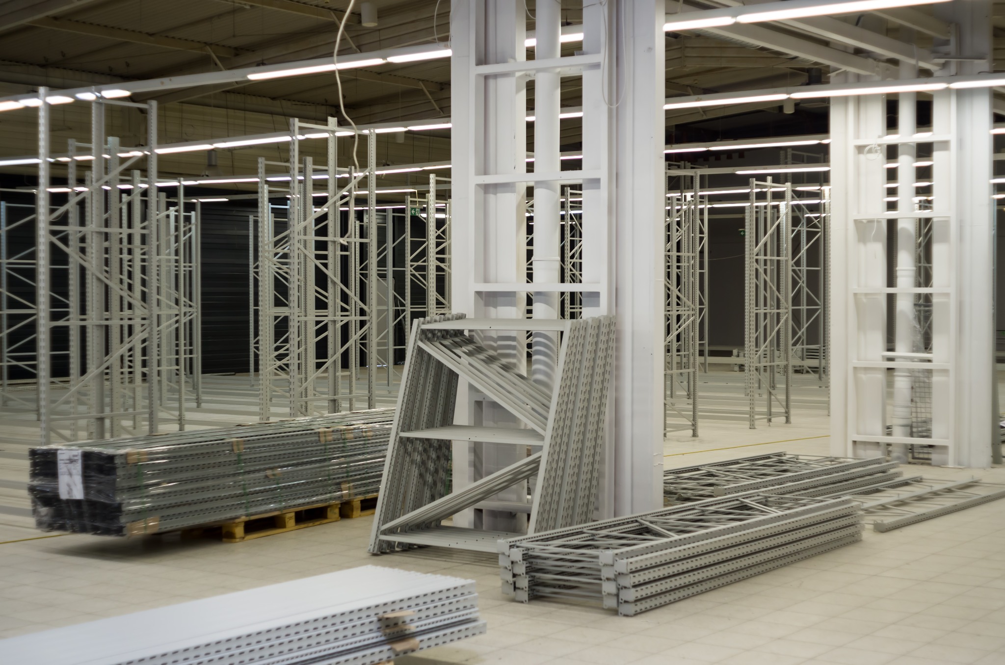 Elementy stalowe konstrukcji magazynowej, przygotowane do montażu, widoczne słupy, belki i poprzeczki, całość w jasnej hali magazynowej.