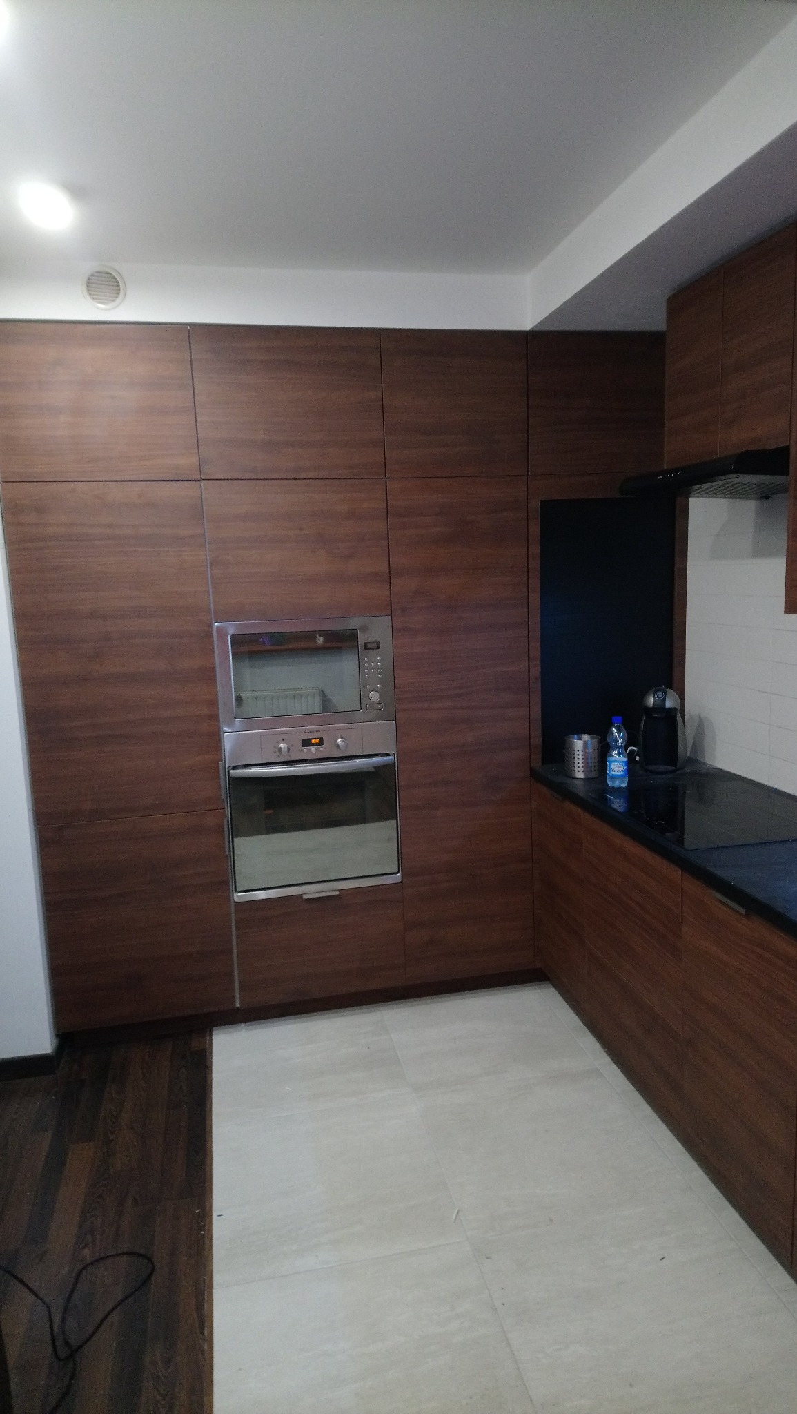 Nowoczesna kuchnia z zabudową w odcieniach ciemnego drewna, z wbudowanym piekarnikiem i czarnym blatem, widok na fragment podłogi z jasnymi płytkami.