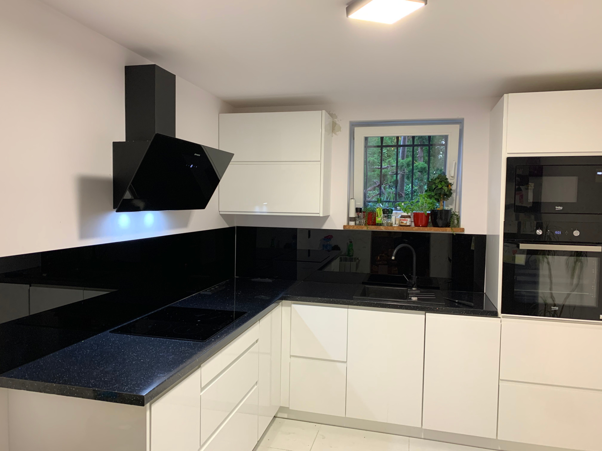 Nowoczesna kuchnia: białe, gładkie szafki, czarny blat z połyskiem, czarny okap, czarny panel nad blatem, okno z widokiem na zieleń.