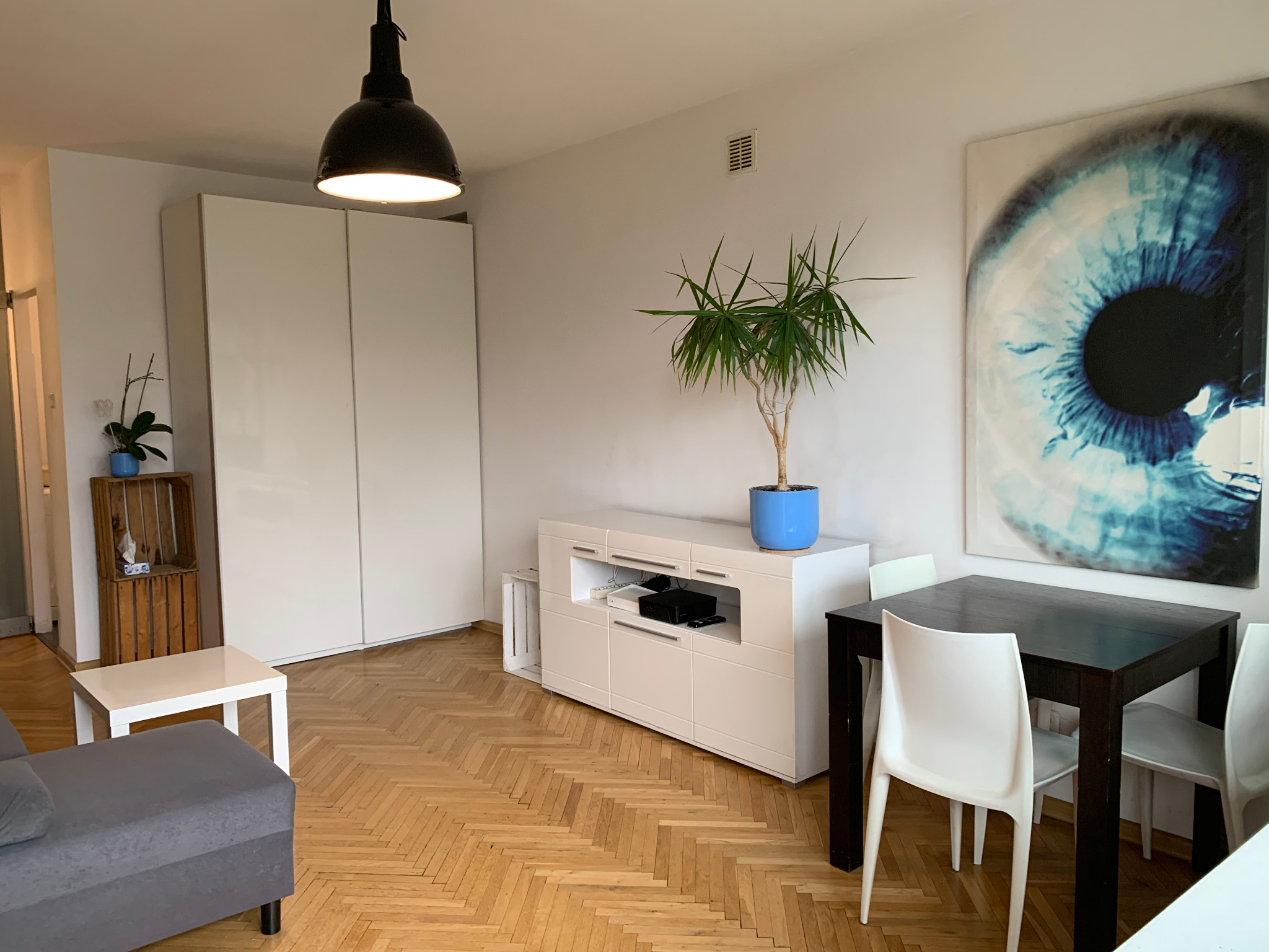 Jasne wnętrze mieszkania z parkietem w jodełkę, białymi meblami, stołem z krzesłami i obrazem oka na ścianie.