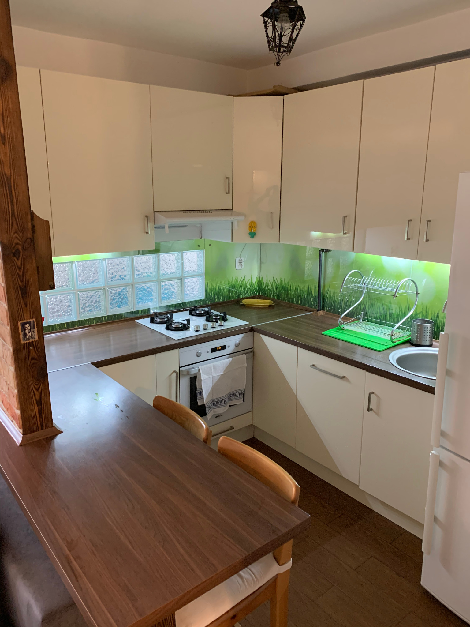 Jasne szafki kuchenne z drewnianym blatem, szklanym panelem z motywem trawy i stołem z drewnianym blatem, oświetlone ozdobną lampą sufitową.