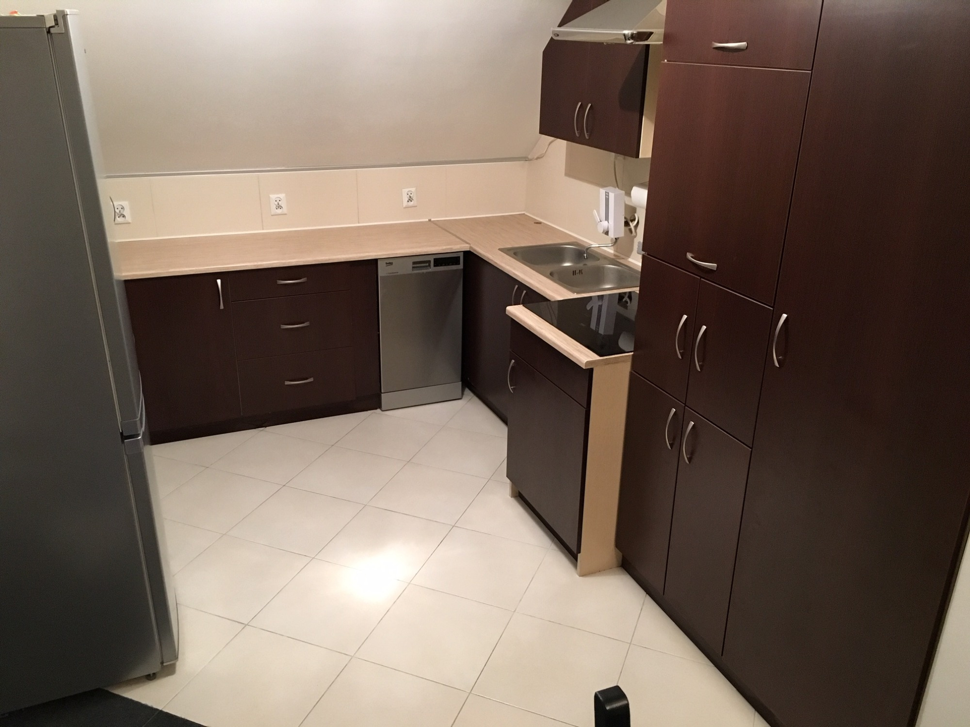 Nowoczesna kuchnia w odcieniach brązu i beżu z białymi kafelkami na podłodze, widoczna zabudowa kuchenna, zlewozmywak, płyta grzewcza i srebrna lodówka.