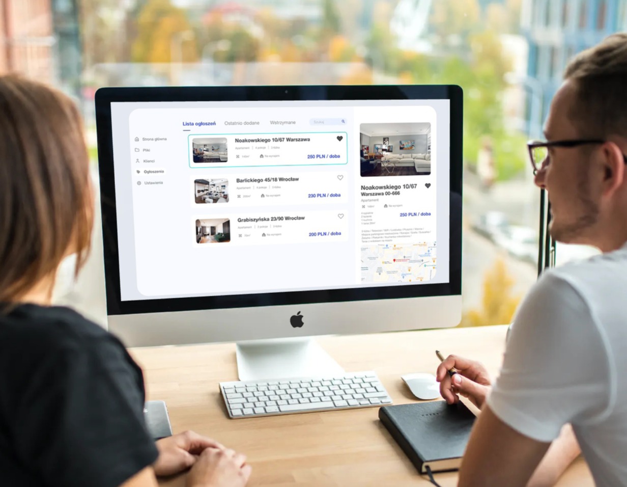Dwie osoby analizują na monitorze komputera listę ogłoszeń nieruchomości z cenami najmu, wyświetlaną w systemie CRM; notatnik i długopis na biurku.