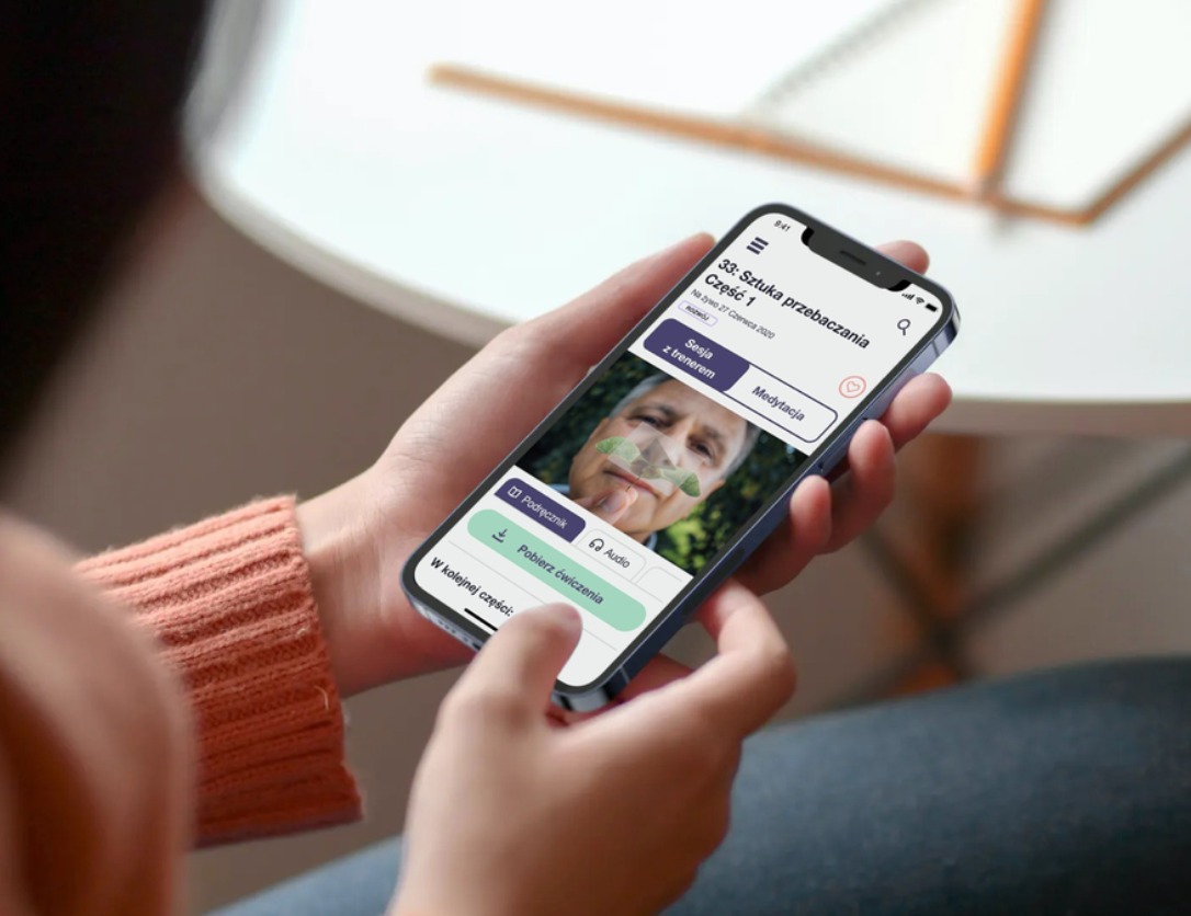 Osoba trzyma iPhone'a z uruchomioną aplikacją do medytacji z sesją z trenerem, widoczny ekran z tytułem '33: Sztuka przebaczania Część 1' i zdjęciem trenera.
