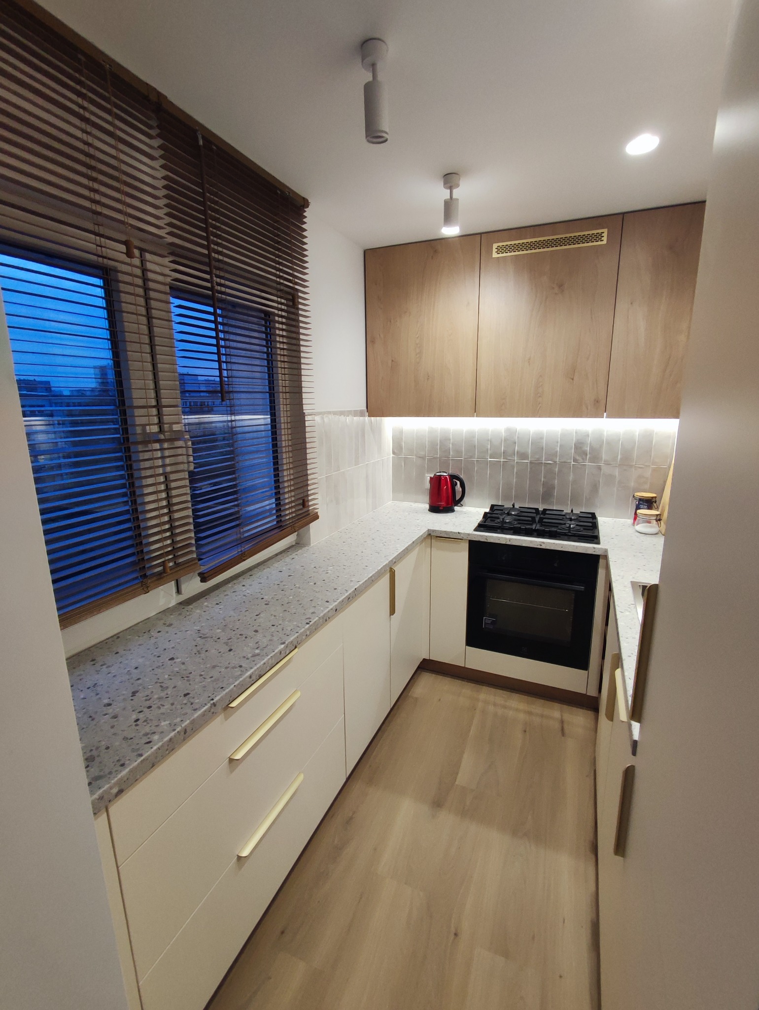 Nowoczesna kuchnia z białymi szafkami z podłużnymi złotymi uchwytami, blatem z szarego lastryko, drewnianymi szafkami wiszącymi i oknem z żaluzjami drewnianymi, na blacie czajnik elektryczny...