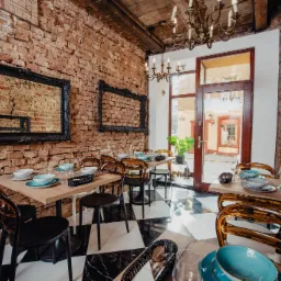 Rustykalne wnętrze restauracji z ceglanymi ścianami, czarno-białą szachownicą na podłodze, drewnianym sufitem i stołami nakrytymi zastawą w odcieniach błękitu i bieli, oświetlone żyrandolem w stylu...