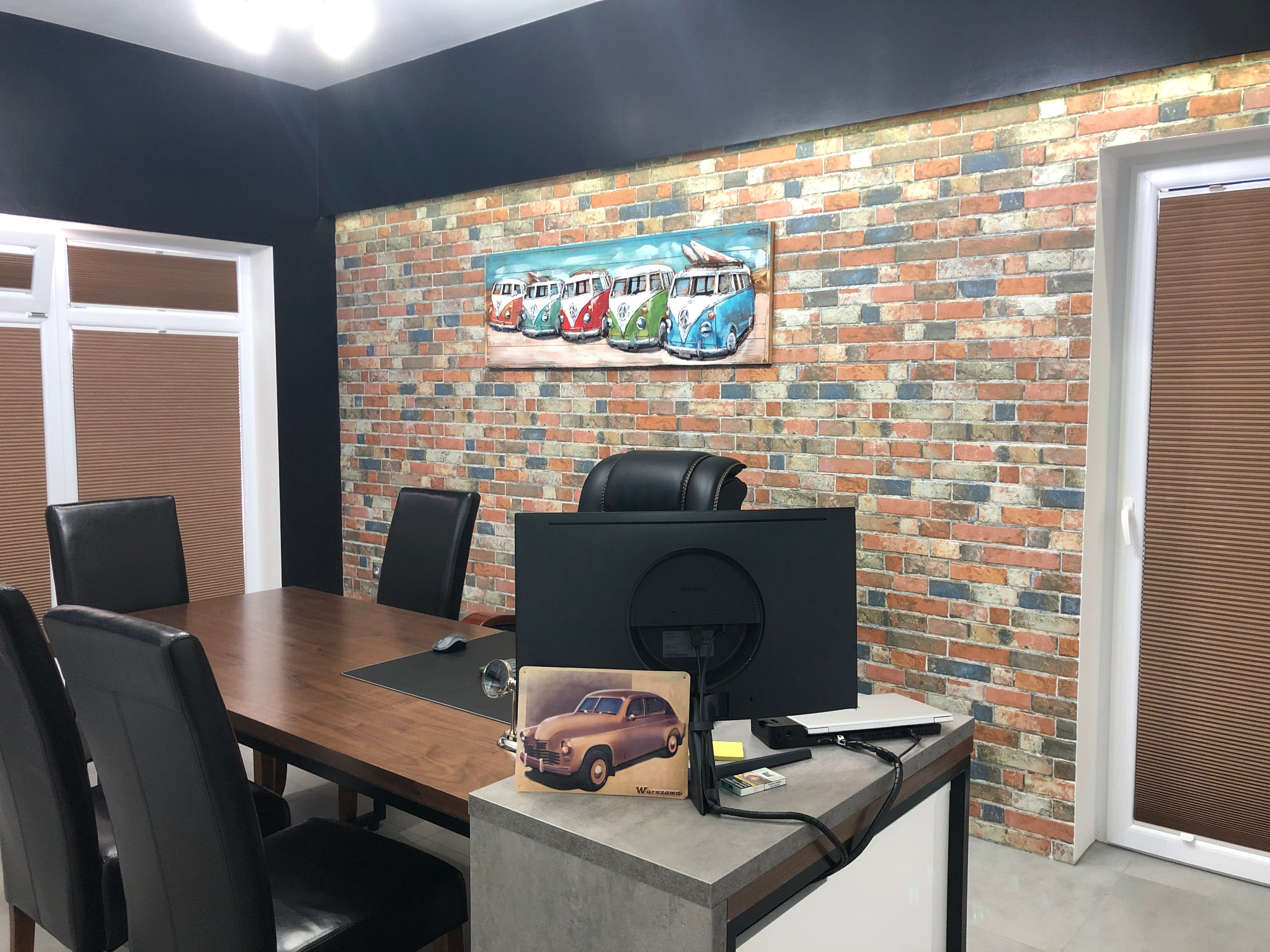 Sala konferencyjna z dużym drewnianym stołem, czarnymi krzesłami i ścianą z cegły, na której wisi obraz z kolorowymi Volkswagenami Transporterami. Na biurku stoi monitor, laptop i metalowa...