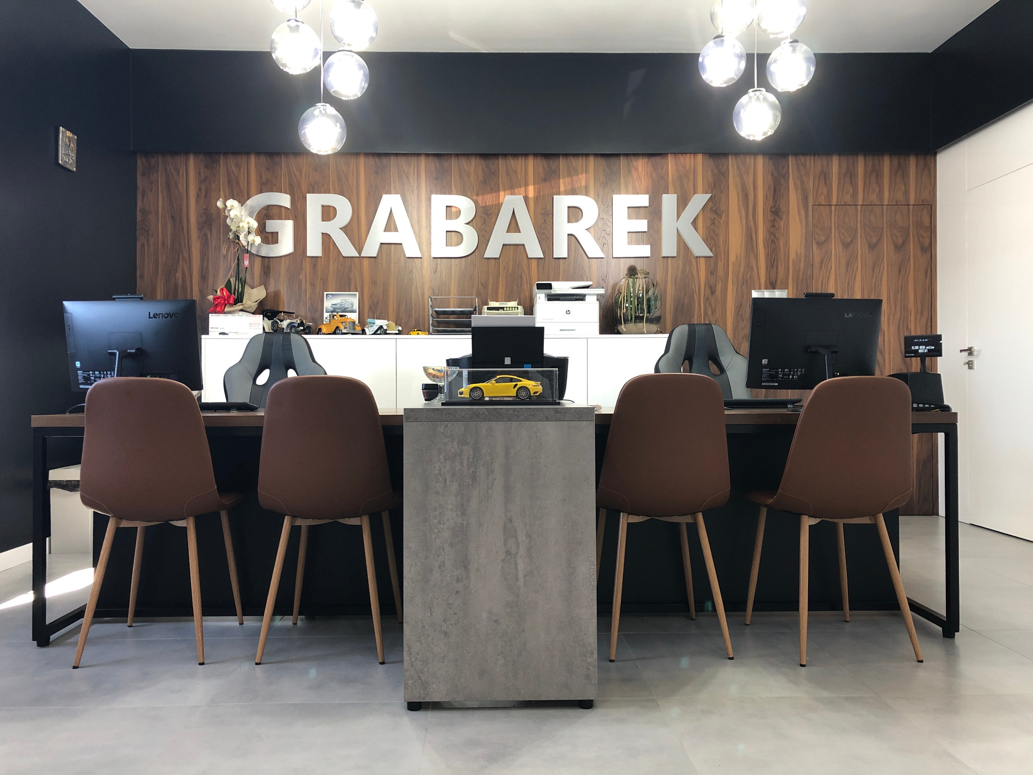 Recepcja warsztatu samochodowego z napisem 'GRABAREK' na drewnianej ścianie, monitory i krzesła w tle, na środku makieta żółtego sportowego auta.