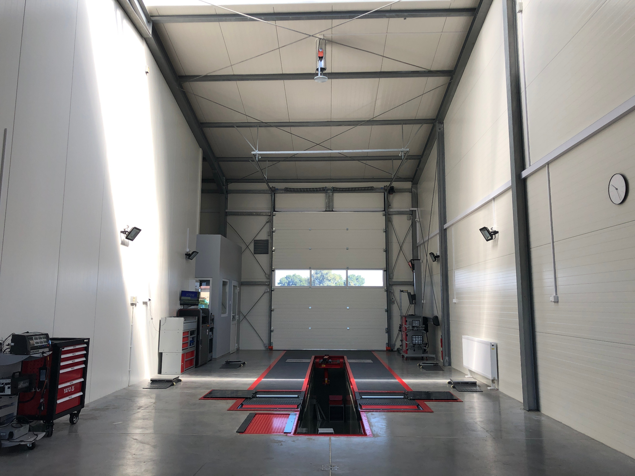 Wnętrze warsztatu samochodowego z podnośnikiem kanałowym, narzędziami i sprzętem diagnostycznym. Widoczne oświetlenie LED, brama garażowa z widokiem na zieleń oraz białe ściany z poziomymi panelami.