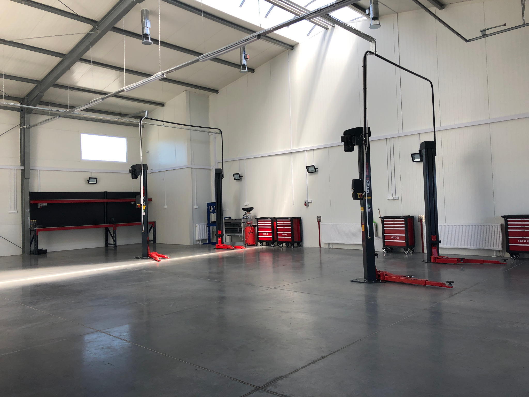 Jasne, przestronne wnętrze warsztatu samochodowego z trzema podnośnikami kolumnowymi, czerwonymi wózkami narzędziowymi Yato i betonową posadzką odbijającą światło słoneczne wpadające przez okno...