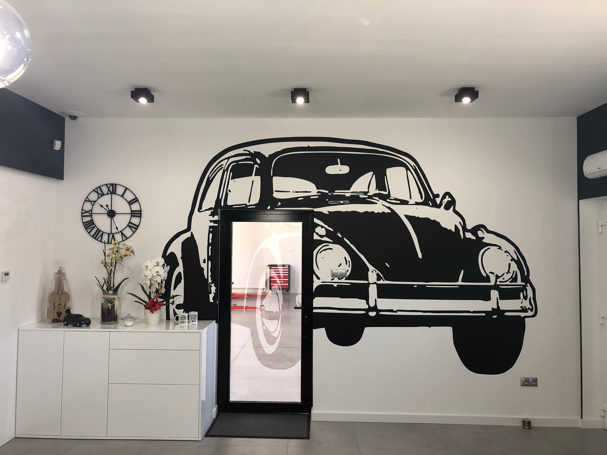 Wnętrze warsztatu samochodowego z dekoracyjnym, czarno-białym malunkiem przedstawiającym Volkswagena Garbusa na ścianie, widoczny przez przeszklone drzwi wjazd na stanowisko z podnośnikiem...