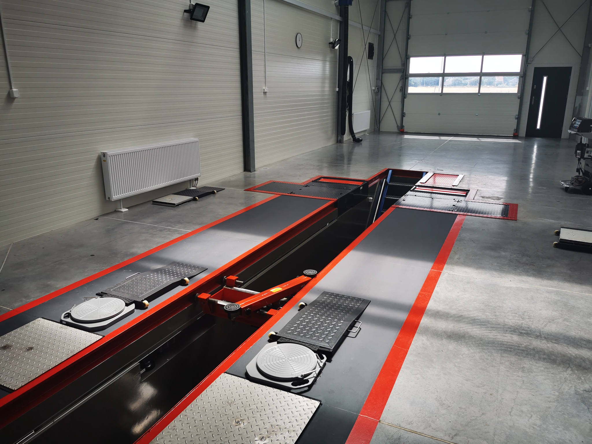 Nowoczesny warsztat samochodowy z kanałem diagnostycznym, widoczne podnośniki i stanowiska do kontroli geometrii kół, hala z białymi ścianami panelowymi i bramą wjazdową.