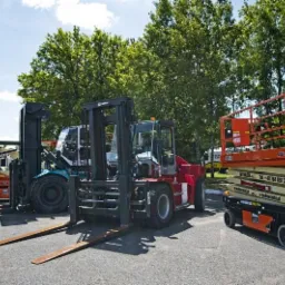 Różnokolorowe wózki widłowe i podnośnik nożycowy zaparkowane na placu, widoczne drzewa w tle.