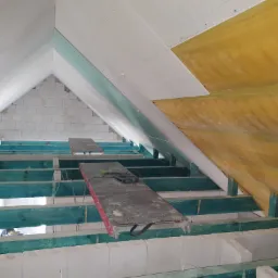 Wnętrze poddasza w trakcie prac: widoczne ocieplenie folią, konstrukcja z belek pomalowanych na niebiesko-zielono, płyty karton-gips częściowo zamontowane, narzędzie na desce roboczej.