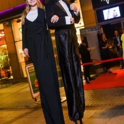 Para performerów na szczudłach w eleganckich, czarnych strojach (cylinder, smoking, damski garnitur) pozuje przed wejściem do klubu nocnego z czerwonym dywanem.