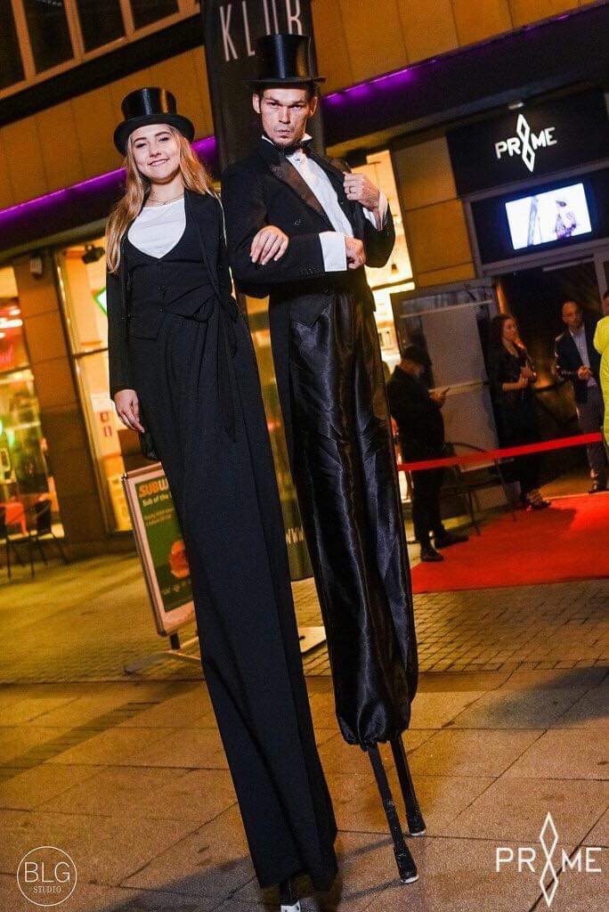 Para performerów na szczudłach w eleganckich, czarnych strojach (cylinder, smoking, damski garnitur) pozuje przed wejściem do klubu nocnego z czerwonym dywanem.