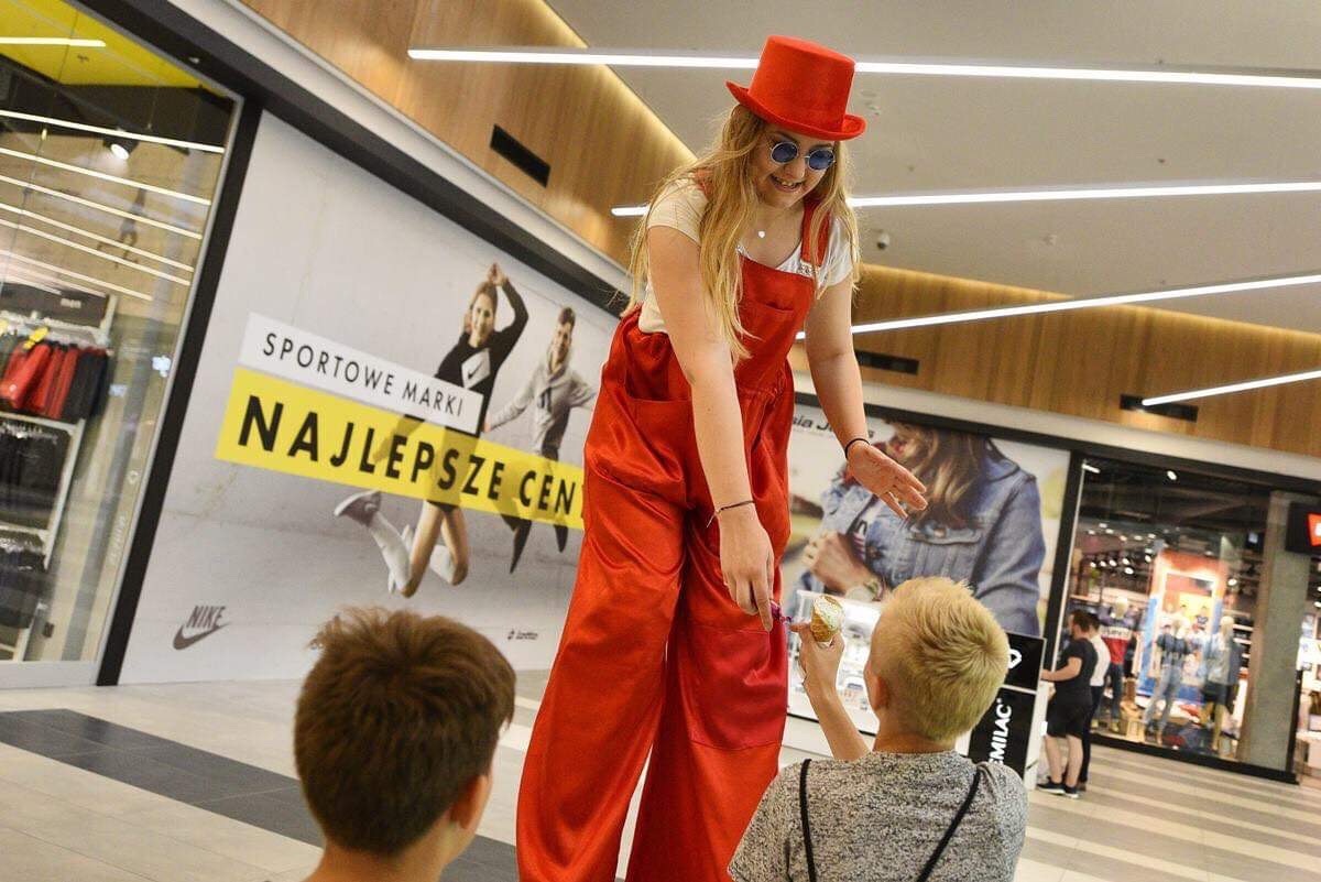 Kobieta w czerwonym kombinezonie i kapeluszu wręcza loda dziecku w centrum handlowym, na tle witryny sklepowej z reklamą sportową.