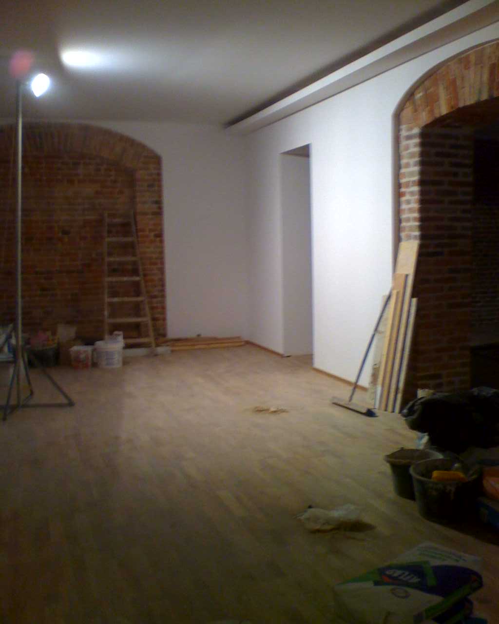 Wnętrze w trakcie prac wykończeniowych: widoczna drewniana podłoga, ceglane łuki, ściany przygotowane do malowania, drabina oparta o ścianę i narzędzia malarskie.