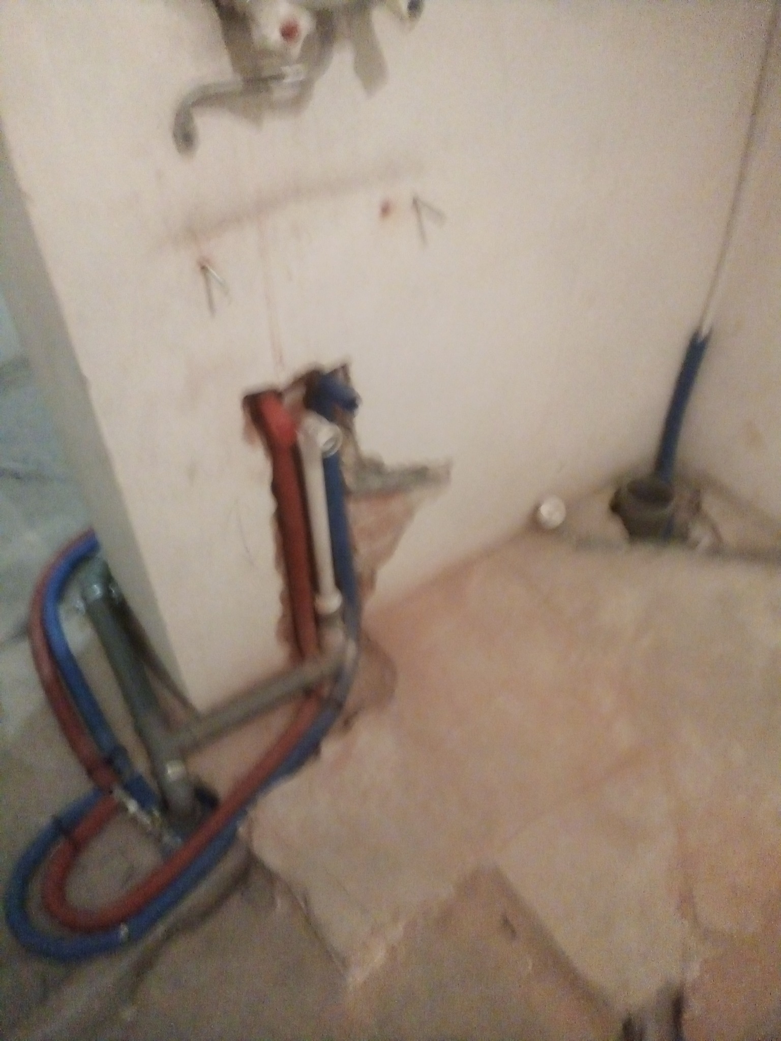 Surowe ściany z wystającymi rurami instalacji wodnej w kolorze niebieskim i czerwonym, widoczne pozostałości po kuciu w ścianie, etap prac instalacyjnych.