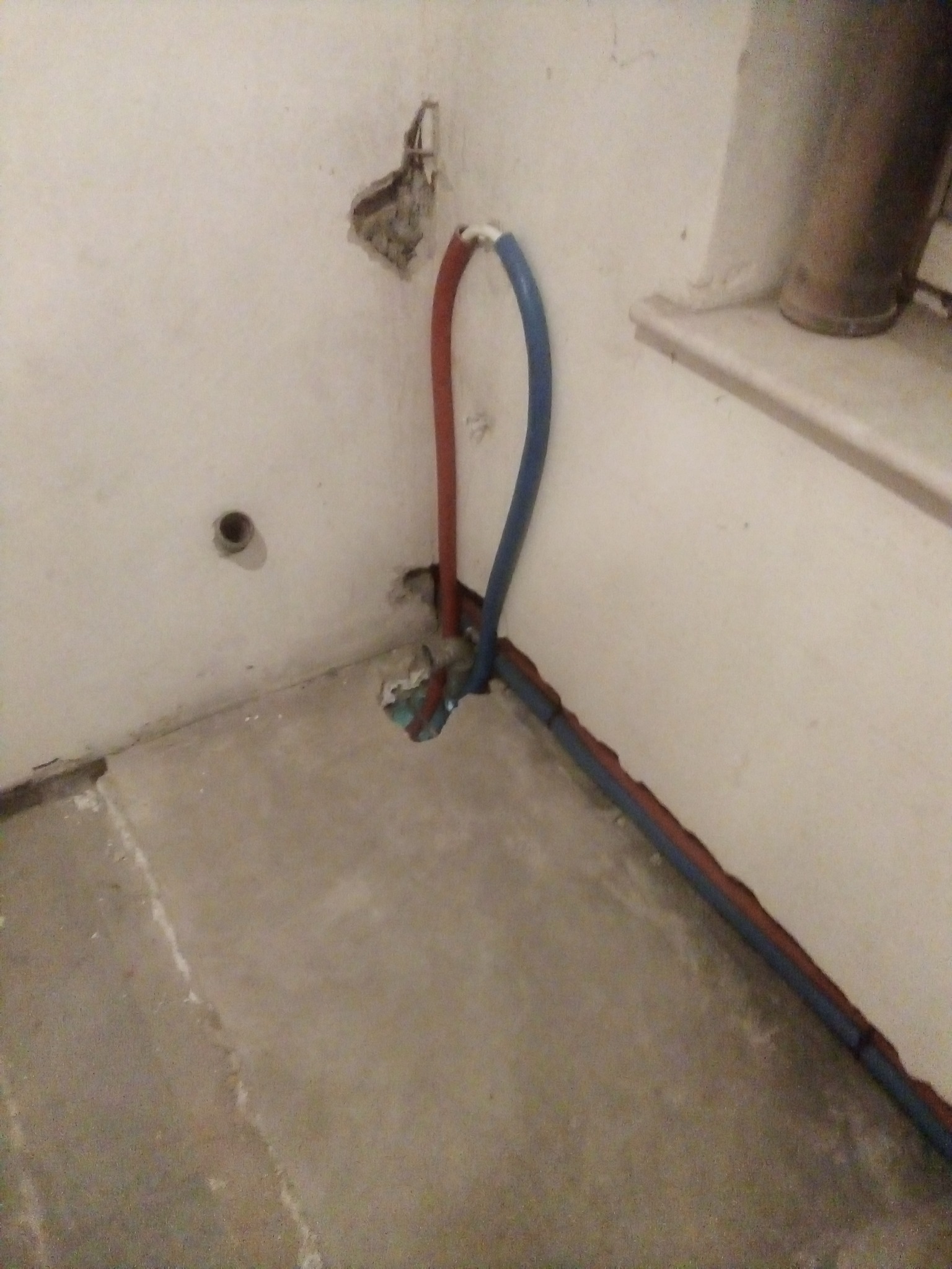Fragment ściany i podłogi w trakcie prac hydraulicznych: widoczne wystające rury w kolorach czerwonym i niebieskim, ślady po kuciu w ścianie, surowy stan wykończenia.