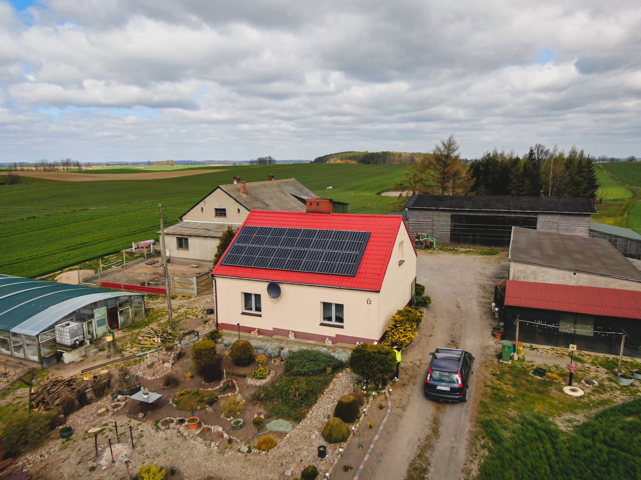 Widok z lotu ptaka na dom z czerwoną dachówką i panelami słonecznymi, otoczony ogrodem z ścieżkami z kamieni, na tle pól uprawnych i pochmurnego nieba.