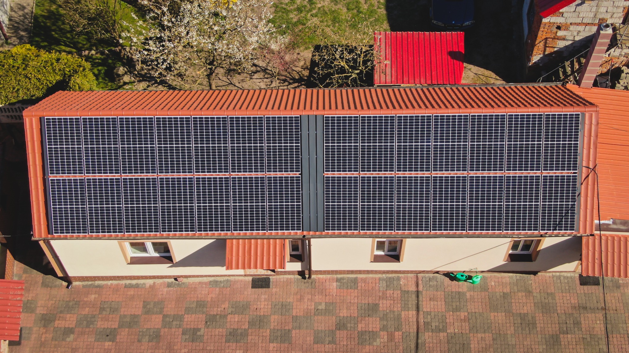 Widok z góry na panele słoneczne na dachu budynku z czerwoną dachówką, obok kwitnące drzewo i czerwony blaszany garaż.