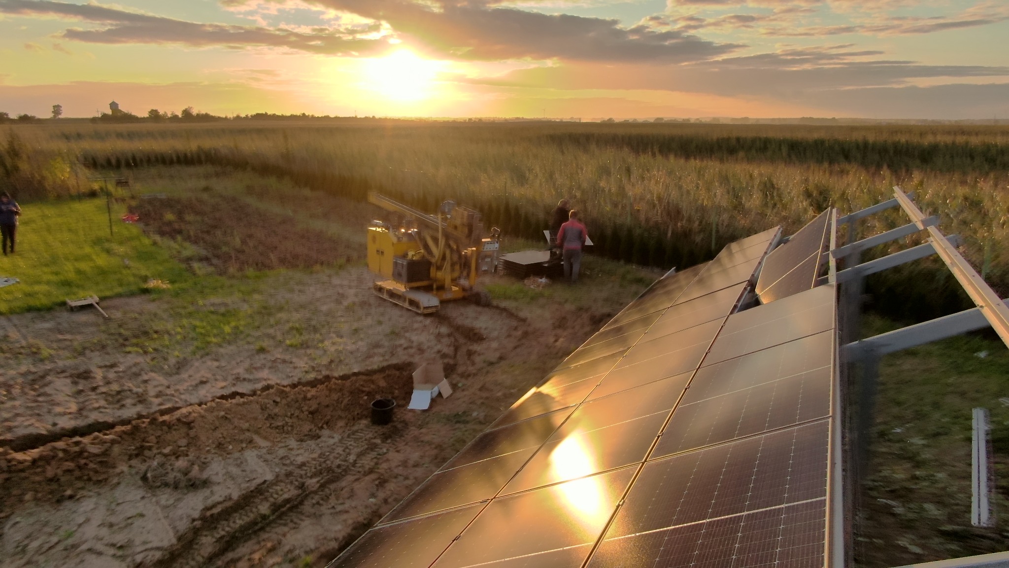 Montaż paneli słonecznych na polu uprawnym o zachodzie słońca, widoczny żółty wiertniczy pojazd i dwóch pracowników.