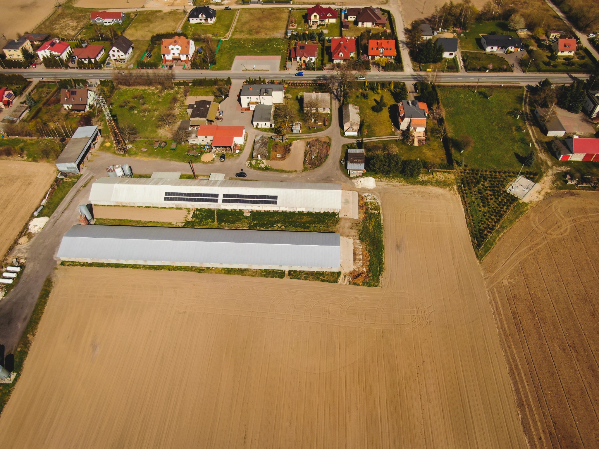 Widok z lotu ptaka na budynki gospodarcze z panelami słonecznymi na dachu, sąsiadujące z polem uprawnym i zabudową jednorodzinną.