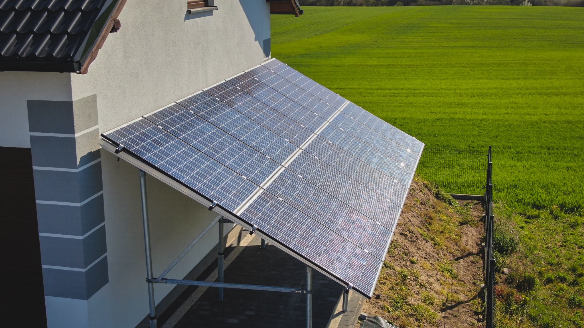 Panele słoneczne zamontowane na wspornikach przy ścianie domu, widok z góry, na tle zielonego pola i ogrodzenia.
