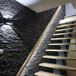 Nowoczesne schody z drewnianymi stopniami i metalowymi wspornikami, oświetlone listwą LED wzdłuż ściany wyłożonej kamieniem dekoracyjnym w odcieniach szarości, z lampą ścienną.