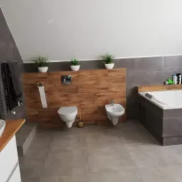 Przestronna łazienka na poddaszu z szarymi płytkami podłogowymi, drewnopodobną ścianką działową, toaletą, bidetem i wanną obudowaną szarą płytką, doświetlona trzema doniczkami z zielonymi roślinami.