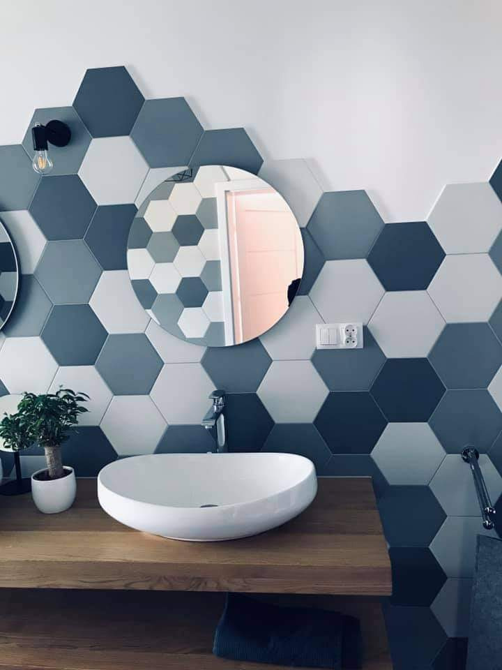 Nowoczesna łazienka z heksagonalnymi płytkami w odcieniach szarości i błękitu, okrągłe lustro nad owalną białą umywalką nablatową na drewnianym blacie, minimalistyczne oświetlenie i roślina...