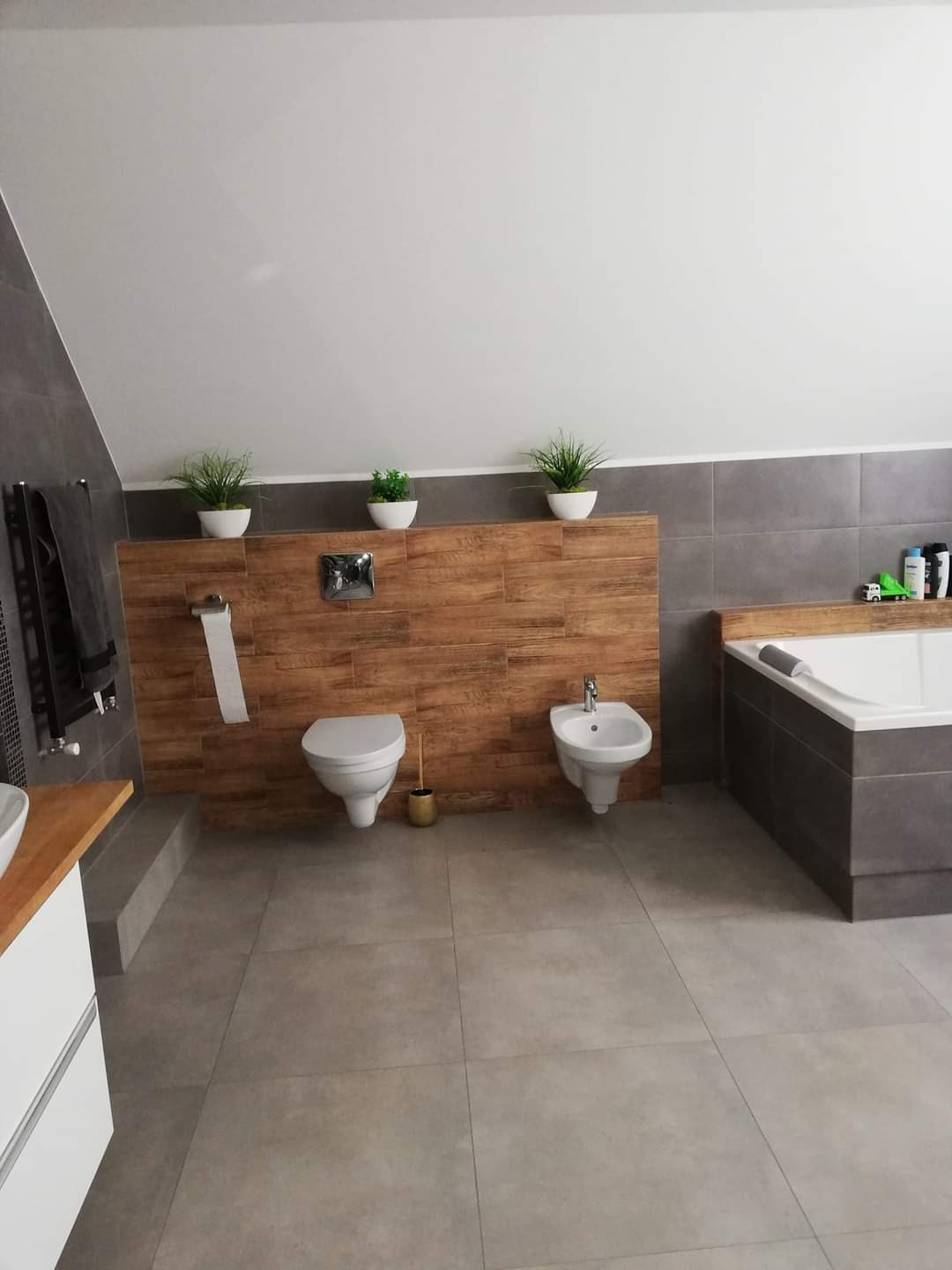 Przestronna łazienka na poddaszu z szarymi płytkami podłogowymi, drewnopodobną ścianką działową, toaletą, bidetem i wanną obudowaną szarą płytką, doświetlona trzema doniczkami z zielonymi roślinami.
