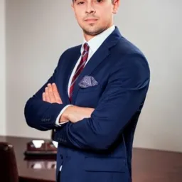 Młody mężczyzna w granatowym garniturze, z poszetką i bordowym krawatem, stoi z założonymi rękami na tle biura z ciemnym drewnianym stołem.