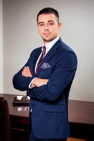 Młody mężczyzna w granatowym garniturze, z poszetką i bordowym krawatem, stoi z założonymi rękami na tle biura z ciemnym drewnianym stołem.