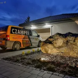 Ż&oacute;łto-szary samoch&oacute;d dostawczy firmy elektrycznej Czamper zaparkowany na podjeździe przed domem z oświetlonym garażem i dekoracyjnym kamieniem z wodospadem w ogrodzie, sfotografowany o zmierzchu.