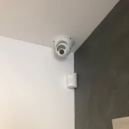 Kamera monitoringu kopułkowa zamontowana na suficie w rogu pomieszczenia, obok czujnik ruchu na ścianie, widoczny fragment ramy drzwi.
