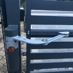 Szary siłownik bramy skrzydłowej zamontowany na słupku i skrzydle bramy, widoczny mechanizm ramienia przegubowego, w tle siatka ogrodzeniowa i fragment chodnika.