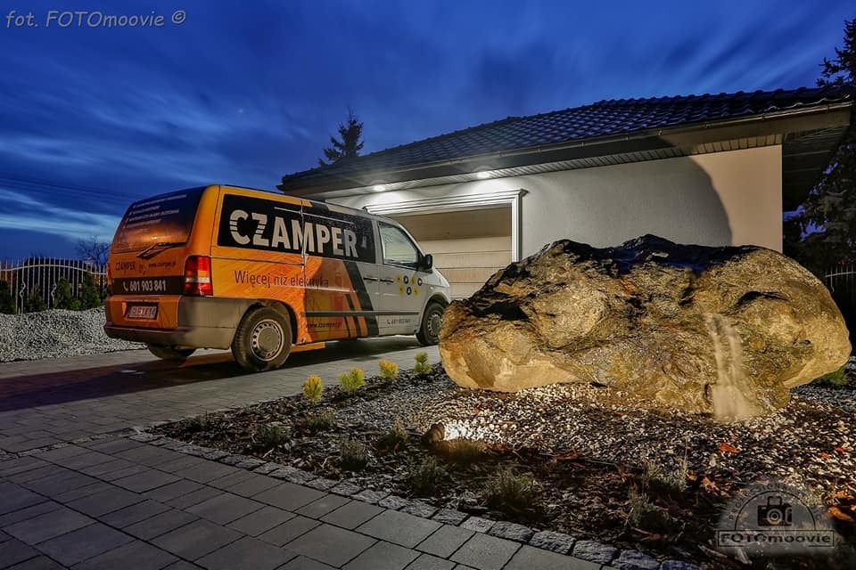 Żółto-szary samochód dostawczy firmy elektrycznej Czamper zaparkowany na podjeździe przed domem z oświetlonym garażem i dekoracyjnym kamieniem z wodospadem w ogrodzie, sfotografowany o zmierzchu.