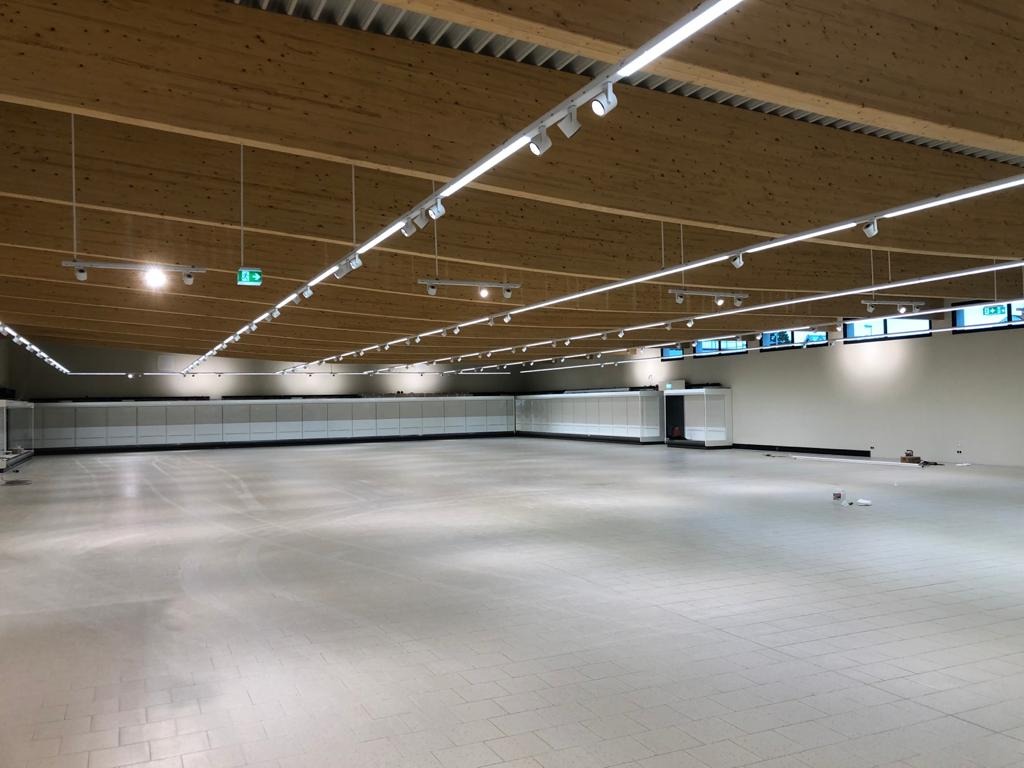 Wnętrze dużego, pustego sklepu z drewnianym sufitem i rzędami lamp sufitowych LED. Widoczne przygotowane półki i instalacja oświetlenia.