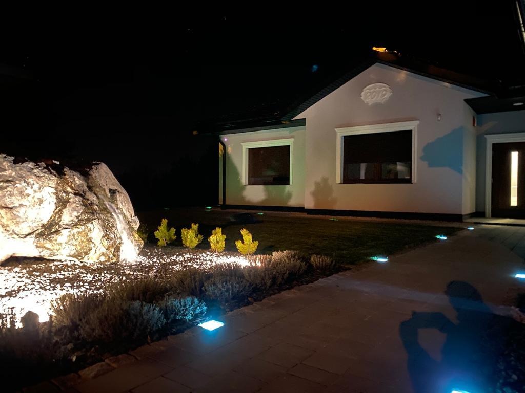 Nocne oświetlenie posesji z podświetlonym kamieniem, roślinami i ścieżką prowadzącą do domu.