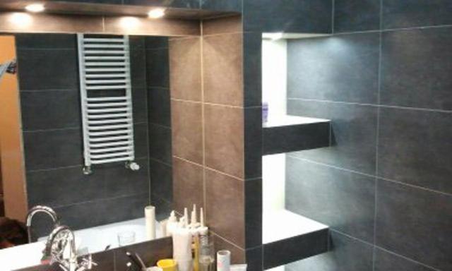 Wykończona łazienka z ciemnymi, kwadratowymi płytkami, grzejnikiem drabinkowym i wnękami z półkami, widok na umywalkę z baterią i akcesoriami.