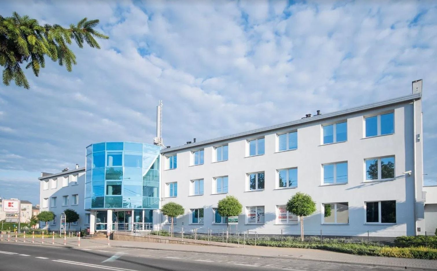 Nowoczesny budynek biurowy z przeszkloną, cylindryczną klatką schodową na rogu, widoczny z ulicy w słoneczny dzień.