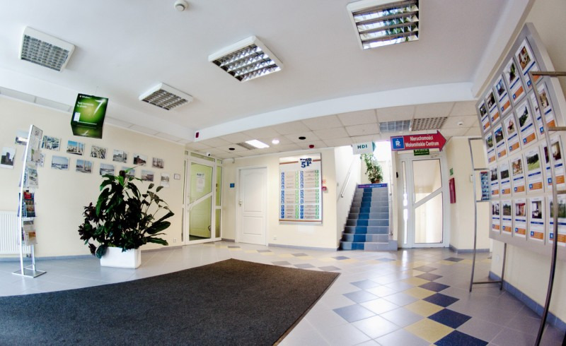 Jasne wnętrze biurowca z rośliną doniczkową, tablicami informacyjnymi i schodami prowadzącymi na wyższe piętro.