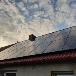 Panele słoneczne na dachu domu jednorodzinnego z czerwonymi dachówkami, widoczne na tle pochmurnego nieba o zachodzie słońca, z kominem i anteną na dachu.